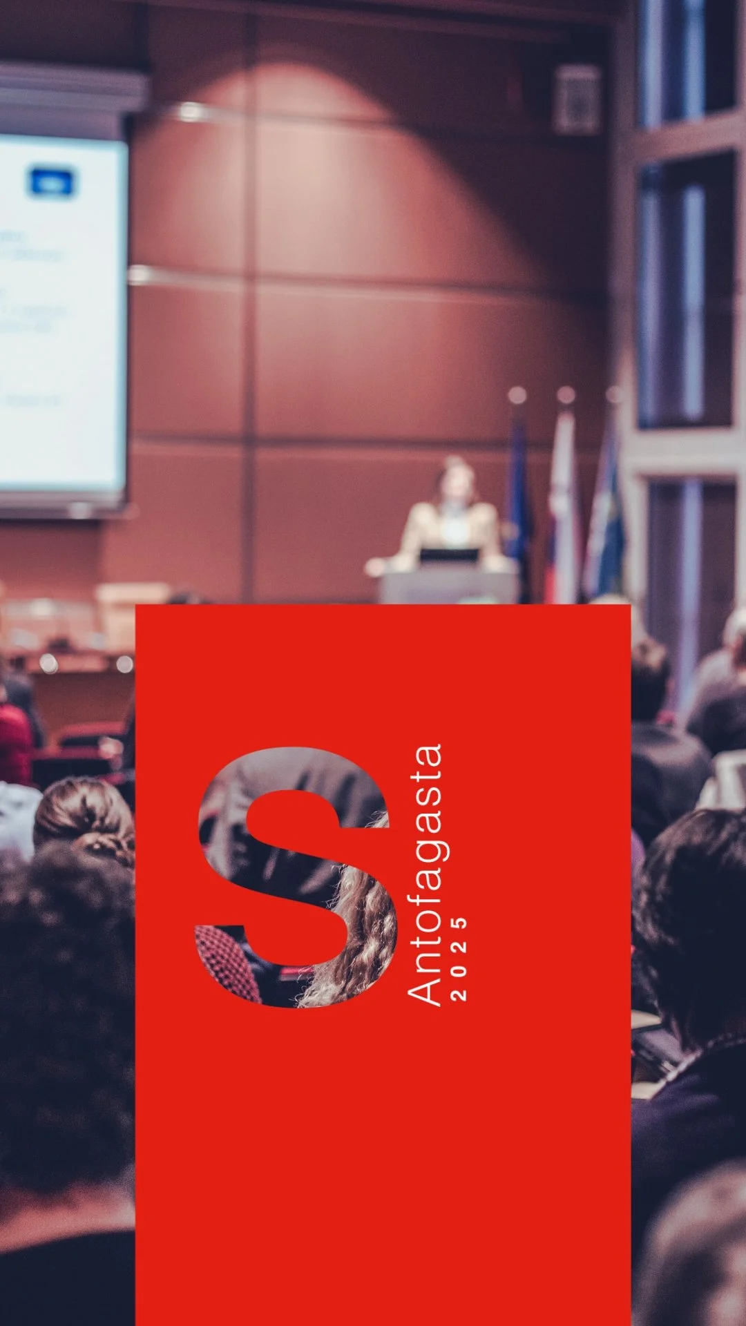 Una conferencia en un auditorio con una pantalla grande y una presentadora en el escenario, asistentes escuchando, cartel en primer plano con texto 'Antofagasta 2025' y un gran 'S' rojo.