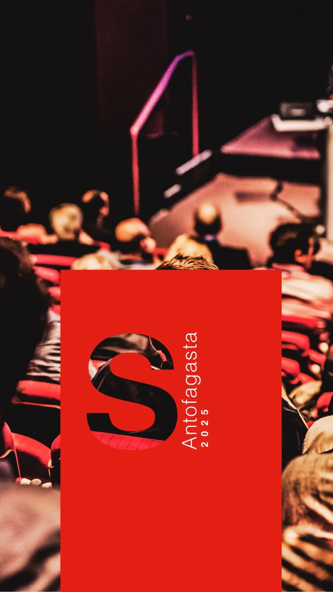 Imagen de un cartel rojo con la palabra 'Antofagasta 2015' y una letra 'S' grande en negro, sobre un fondo de un auditorio con personas y un escenario.